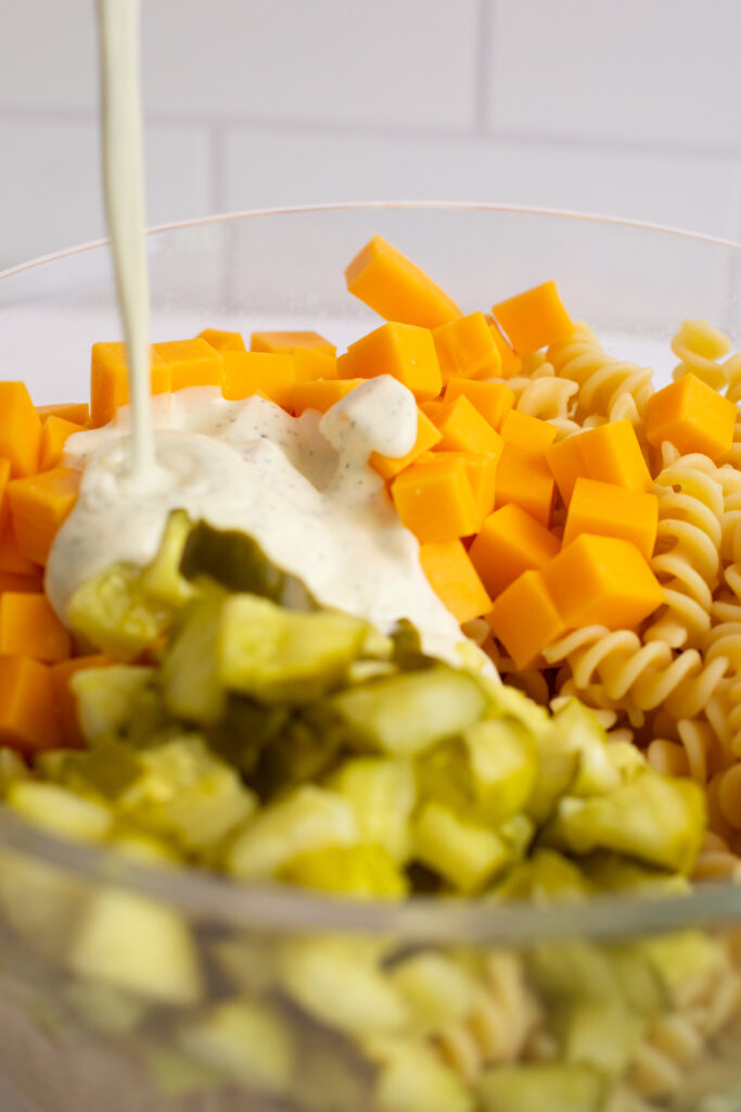 dressing being poured over cheese and pickles and pasta