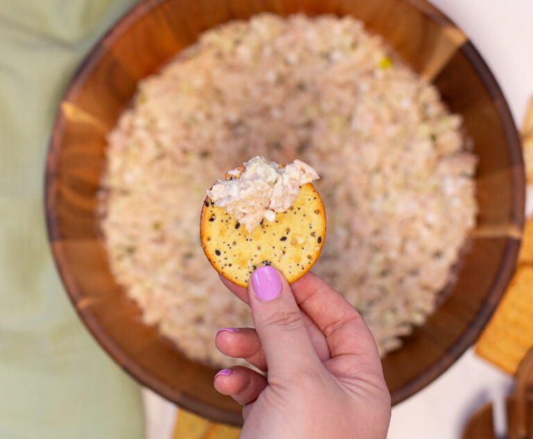 womans hand holding a cracker with some tuna salad over the tuna salad mixing bowl