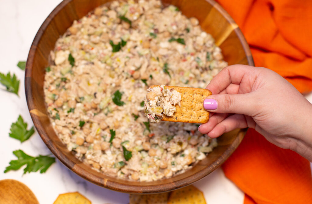 woman's hand holding a cracker with tuna salad on the cracker over a bowl filled with salad