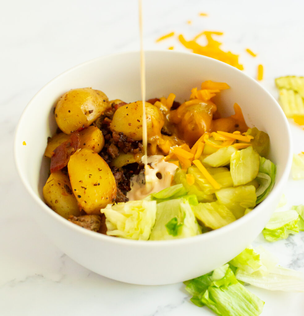 sauce being poured over beef bacon and potato mixture with pickles, lettuce and cheese