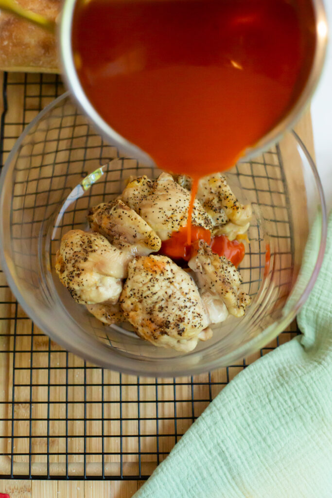 hot sauce being poured over chicken thighs in a bowl