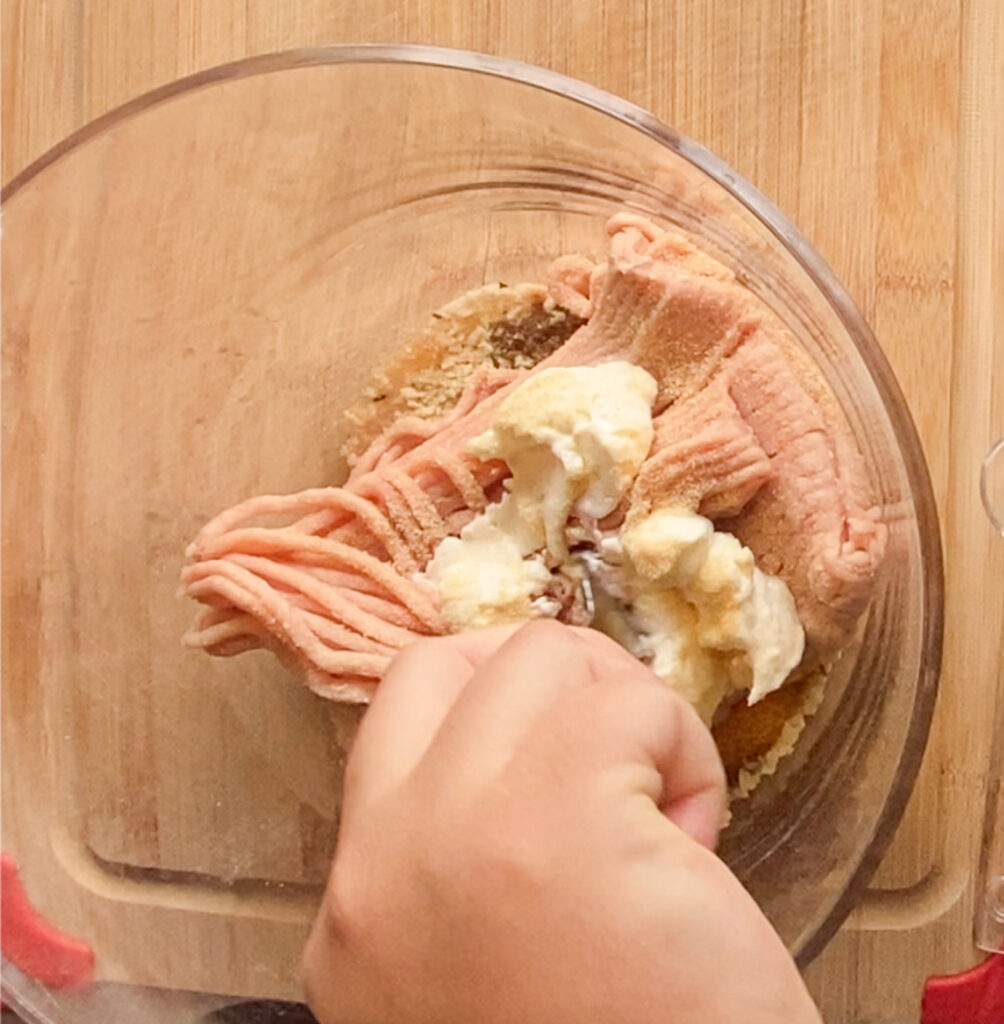 woman's hand mixing together ground chicken, mayo, bread crumbs, and seasonings together in a bowl on top of a cutting board