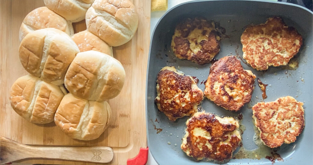 cooked chicken burgers in a skillet with buns to the left of the picture on a cutting board