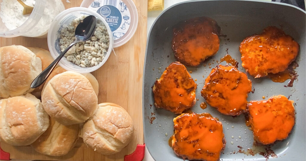 buns and blue cheese on a cutting board to the left of the picture and on the right is chicken burgers covered in buffalo sauce in a skillet
