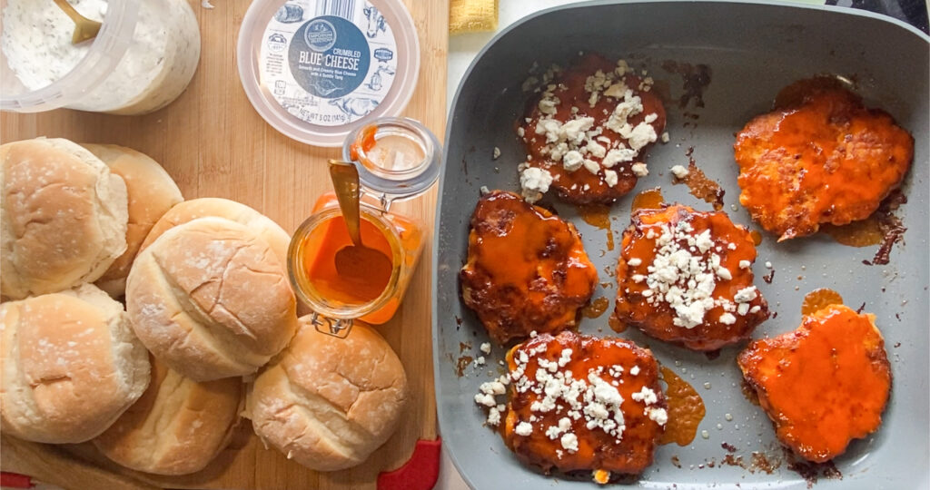 buns, buffalo, and ranch on a cutting board and cooked buffalo chicken burgers with blue cheese in the skillet on the right side of the picture