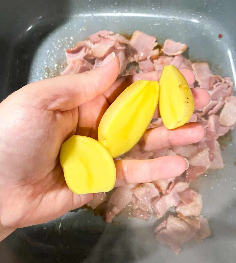 a woman's hand holding halved baby yellow potatoes over the skillet of cooking bacon