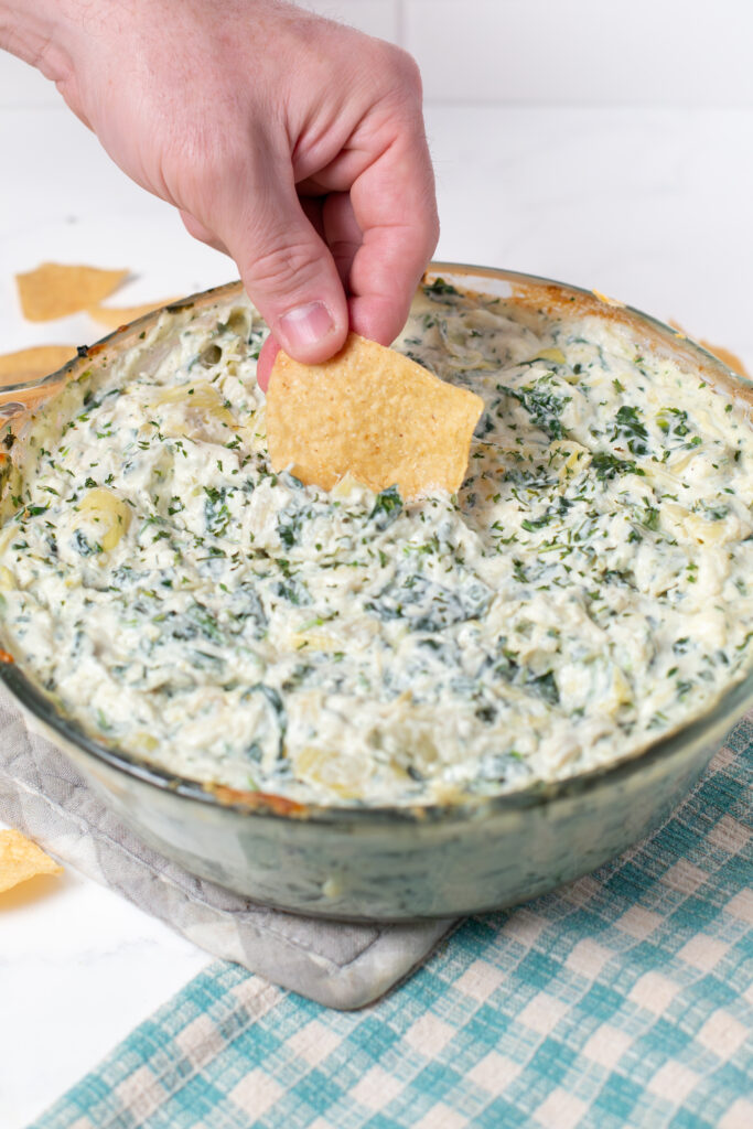a man's hand dipping a tortilla chip in a bowl of spinach artichoke dip