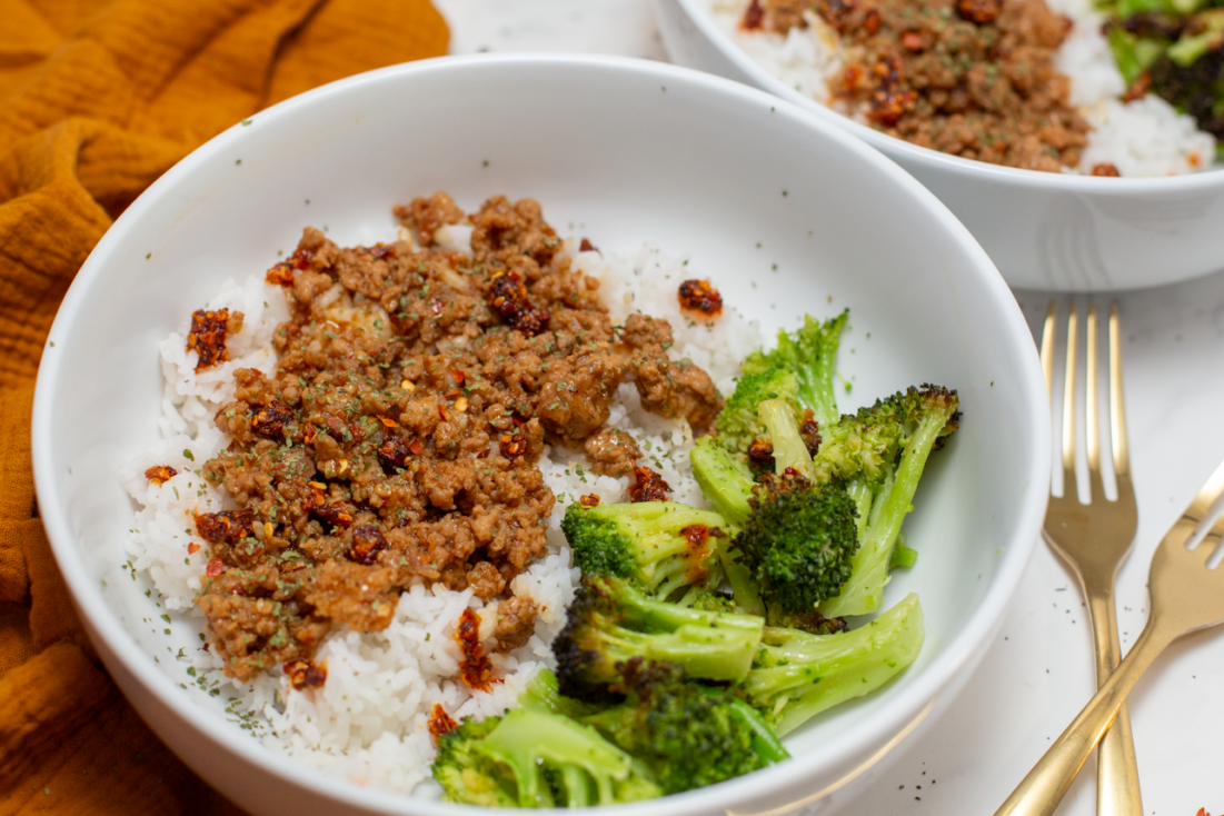 easy asian pork and rice in a white bowl with broccoli on a white counter with gold forks next to it