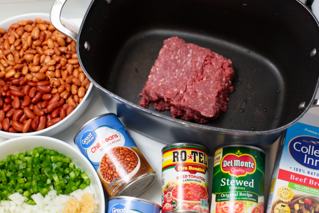 a pot of raw meat, a bowl of beans, a bowl of chopped veggies, canned chili beans, canned tomatoes, and broth on a white counter