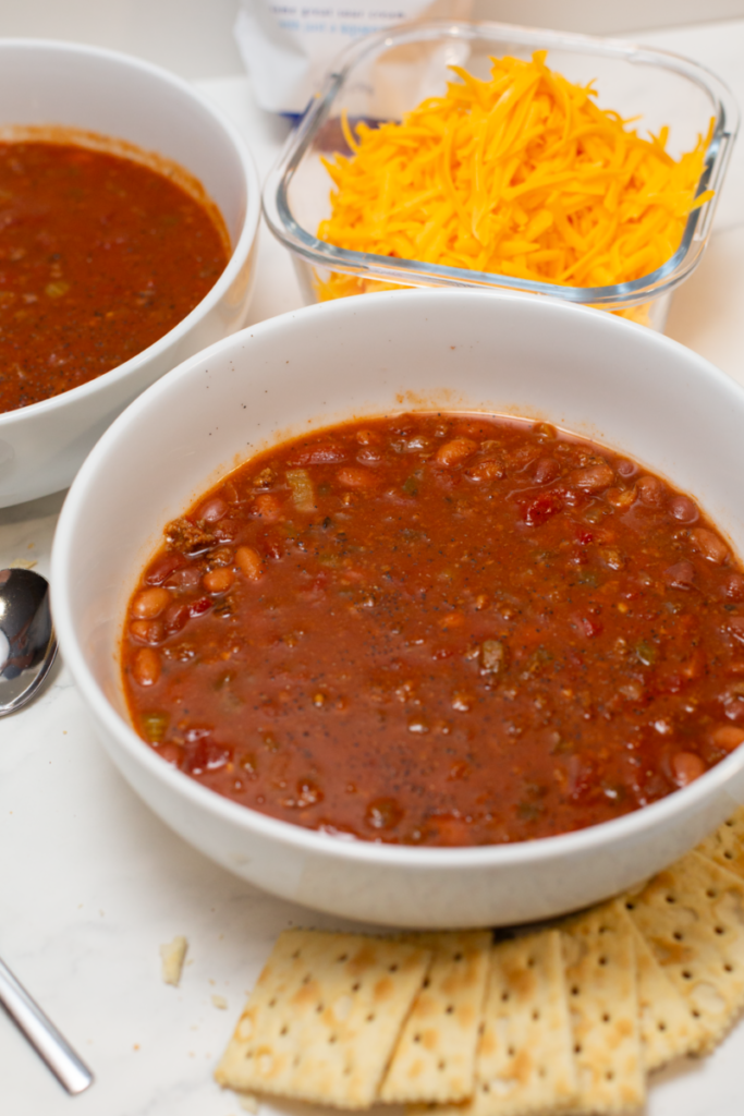 a white bowl full of chili on a white counter surrounded by crackers and a bowl of shredded cheddar