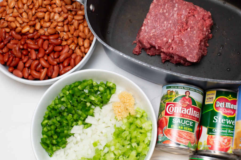 ground meat in the slow cooker, a bowl of beans, a bowl of chopped vegies, and cans of tomato sauce on a white counter