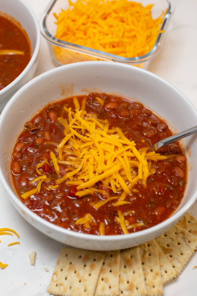 bowl of chili in a white bowl on a white counter with crackers surrounding, a spoon in the bowl, and shredded cheese on top