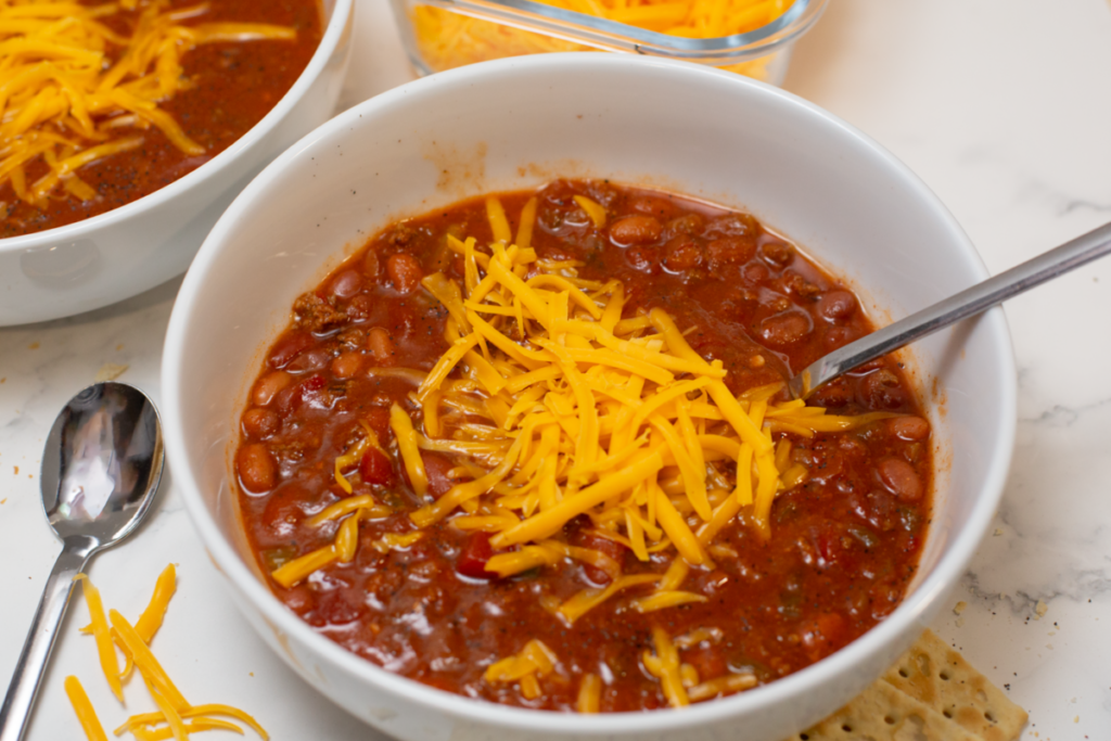 a white bowl of chili topped with cheddar cheese with a spoon resting in the bowl on a white counter surrounded by crackers