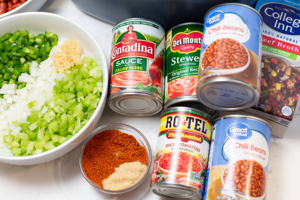 cans of beans and tomato sauce, brother, a bowl of chopped veggies, and a bowl of seasoning on a white counter