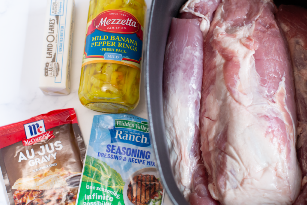 the ingredients for mississippi pork roast on a counter - stick of butter, jar of banana peppers, au just packet, ranch packet, and a pork loin sitting in a slow cooker