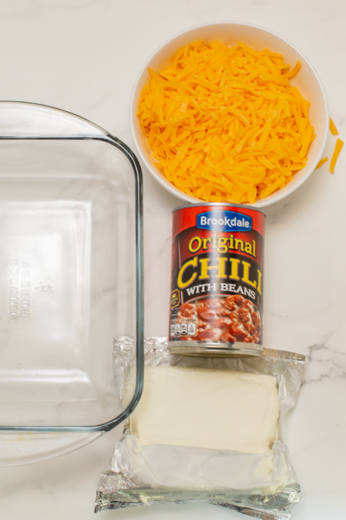 ingredients for the chili dip - shredded cheddar cheese, canned chili, a block of cream cheese, and a glass casserole dish to the side on a white counter