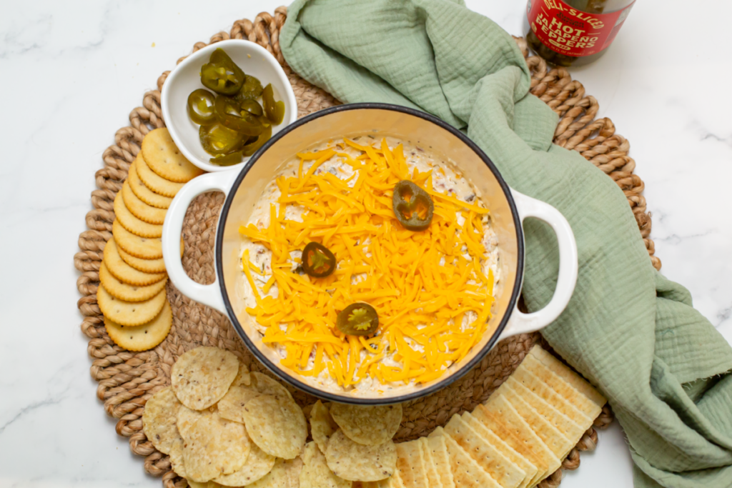 the creamy cheddar jalapeno bacon ranch dip surrounded by crackers and chips and a bowl of pickled jalapenos on a brown tray