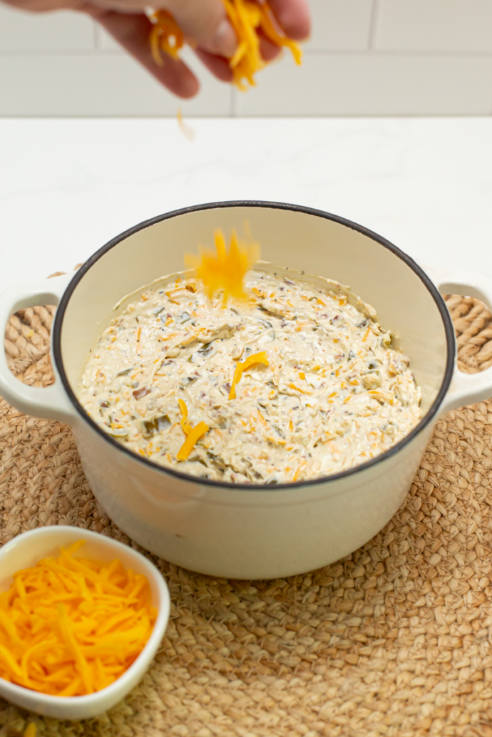 the serving crock with the creamy cheddar jalapeno bacon ranch dip in it and being topped with shredded cheese