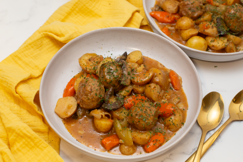 a bowl of meatball stew on a white counter with spoons laying next to it