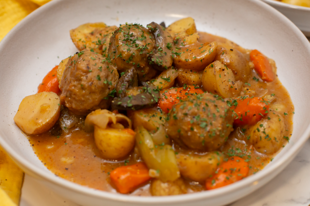 A bowl of meatball stew with a yellow hand towel next to it and a spoon on the other side of it. 